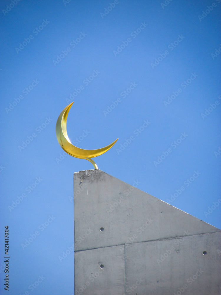 Naklejka premium Crescent moon on top of a modern mosque minaret against the vivid blue sky. A minaret is a visual focal point of a mosque and also provides vantage point to perform the call to prayer 5 times a day.