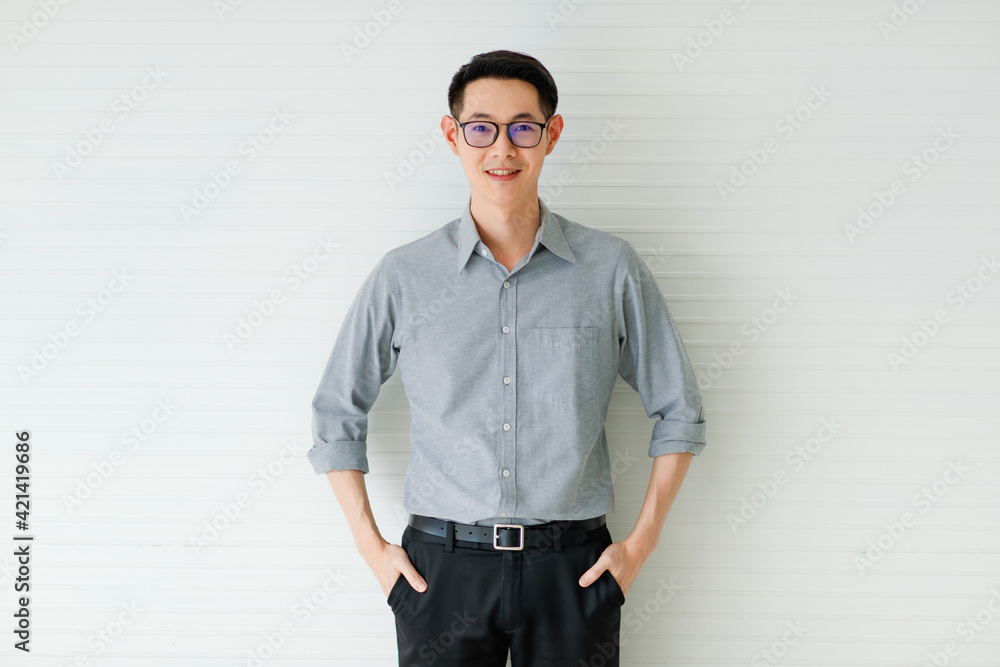 Medium portrait of a formal Asian handsome man with cute smiling, wearing eyeglasses in a gray long sleeve shirt and black pants with hand in pocket posing on white background in the studio Medium portrait of a formal Asian handsome man with cute smiling, wearing eyeglasses in a gray long sleeve shirt and black pants with hand in pocket posing on white background in the studio
