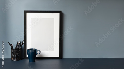 Fototapeta Naklejka Na Ścianę i Meble -  Contemporary workplace with empty frame, coffee cup and copy space on dark blue table.