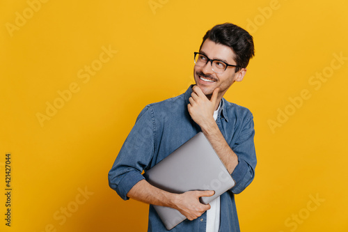 Attractive confident smart guy in glasses and in a denim shirt, holding a laptop at hand, looks thoughtfully towards empty space aside touching his chin, smiling, isolated orange background,copy space