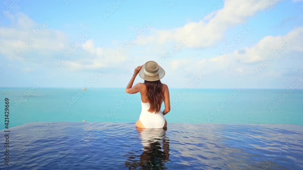 Sitting on the invisible edge of an infinity-edge pool a young woman with her back to the camera greets the world with open arms. Sitting on the invisible edge of an infinity-edge pool a young woman with her back to the camera greets the world with open arms.