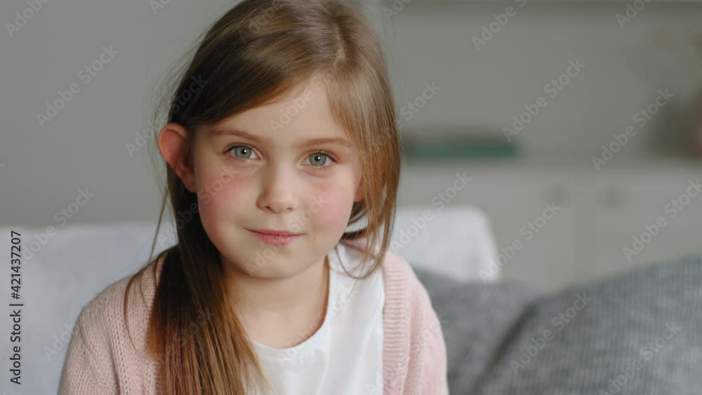 Child portrait, close-up calm friendly face of little child kid, cute baby preschool girl pupil sitting on sofa at home indoor, looking at camera smiling, concept of innocence and happy childhood Child portrait, close-up calm friendly face of little child kid, cute baby preschool girl pupil sitting on sofa at home indoor, looking at camera smiling, concept of innocence and happy childhood