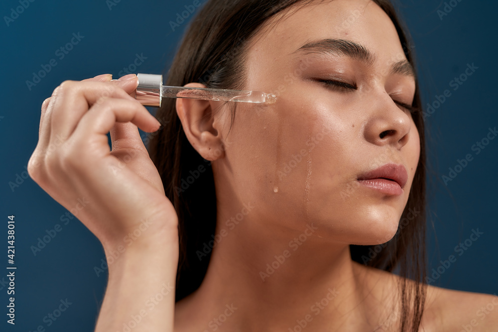 Obraz premium Beautiful asian woman keeping eyes closed applying drops for face care from a pipette on the cheek while standing against dark blue background