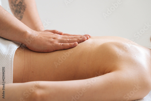 Side view of male masseur massaging small of back of young woman lying on massage table at light spa salon. Experienced chiropractor performs wellness treatments for lady with back pain.