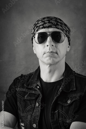 Unshaven, middle-aged man in a T-shirt, denim vest, bandana and sunglasses. Black and white portrait. Photo taken in studio