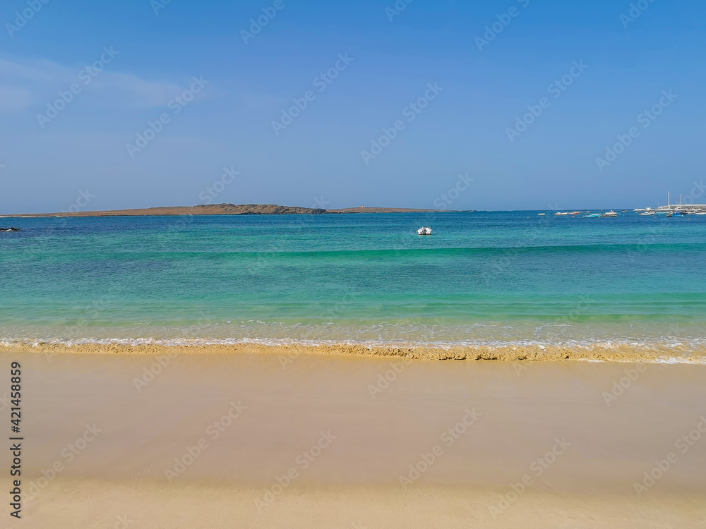 The marine of Sal Rei, Cape Verde. Sandy beach and warm blue water of Atlantic Ocean in the morning sunlight. Selective focus on the waves, blurred background.