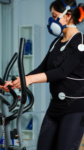 Medical researcher using laptop while measuring sportsman endurance using body sensors, electrodes and mask measuring cardiac rhythm. Patient running on cross trainer in sports science lab photo