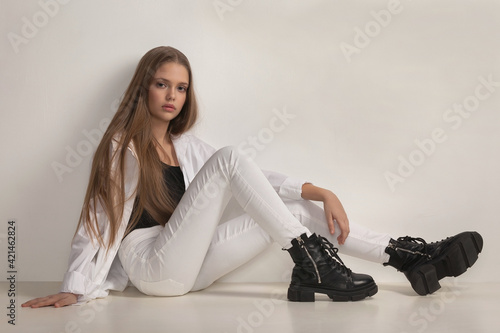 beautiful girl teenager with stylish transparent makeup in white jeans, shirt and boots posing while sitting in the studio on a white background