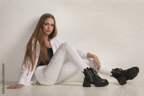 beautiful girl teenager with stylish transparent makeup in white jeans, shirt and boots posing while sitting in the studio on a white background photo