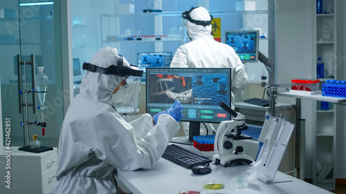 Fotografia Chemist doctor in ppe suit working at pc while lab technician bringing her blood samples
