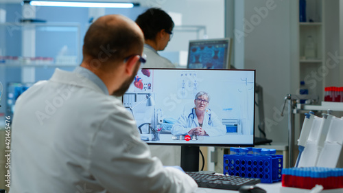 Senior woman doctor offering medical online advices to chemist using pc webcam. Scientist holding blood sample during online discussion  virtual conference  helping on telemedicine  healthcare support