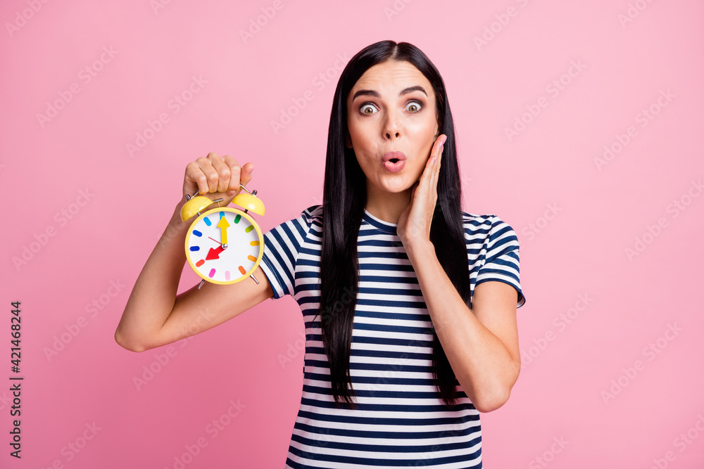 Photo portrait of shocked woman touching cheek face holding yellow clock in one hand isolated on pastel pink colored background Photo portrait of shocked woman touching cheek face holding yellow clock in one hand isolated on pastel pink colored background