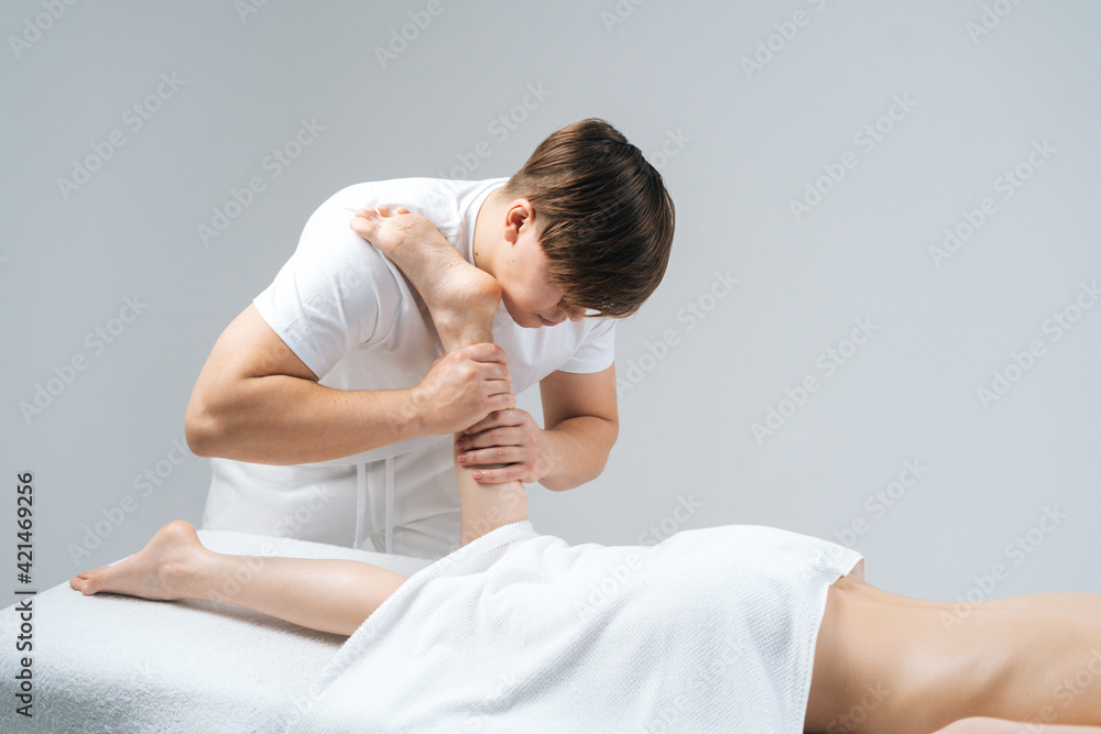Male masseur with strong hands massaging lower part of leg to young woman lying on massage table on white background. Professional therapist men doing wellness massage shins and legs for female client Male masseur with strong hands massaging lower part of leg to young woman lying on massage table on white background. Professional therapist men doing wellness massage shins and legs for female client