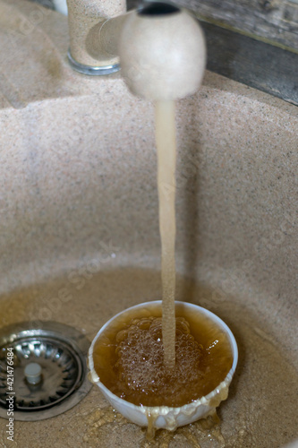 Dirty rusty water flowing from the mains. Faucet with bad water in the house in the kitchen. orange-brown water is collected in a white cup. Poor ecology, polluted environment, lack of clean water. photo