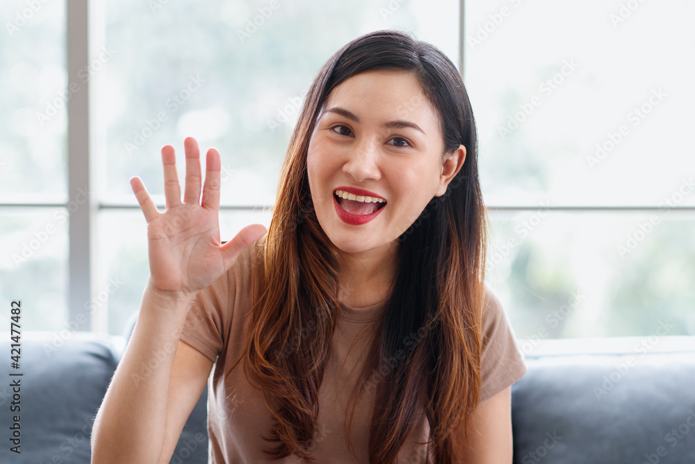 Close up portrait head shot of Asian long black and brown hair young beautiful female wearing brown shirt sitting on blue sofa smiling hold hand up to say hi happily in front glass windows background