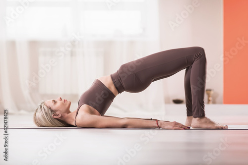 Attractive blonde woman in sports beige leggings and tank top, practicing yoga, doing Glute bridge exercise, dvi pada pithasana pose, home workout