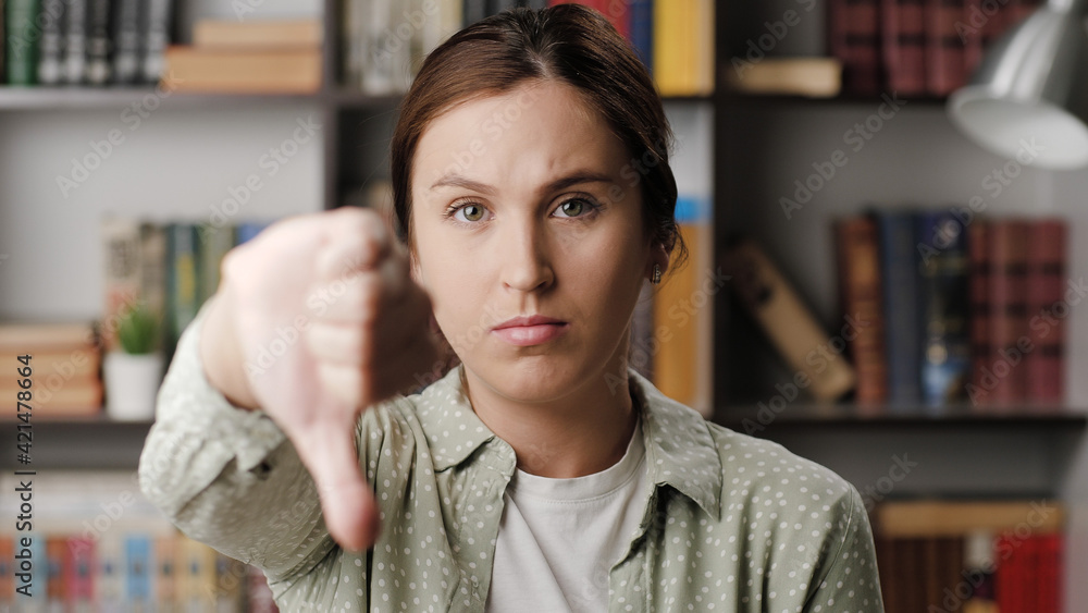 Fototapeta premium Woman thumb down. Frowning serious woman in office or apartment room looking at camera raises her hand and shows her thumb down. Medium shot