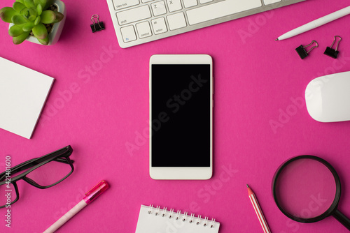 Top view photo of workplace with smartphone in the middle keyboard mouse chancellery magnifier plant glasses and notebooks on isolated pink background with copyspace