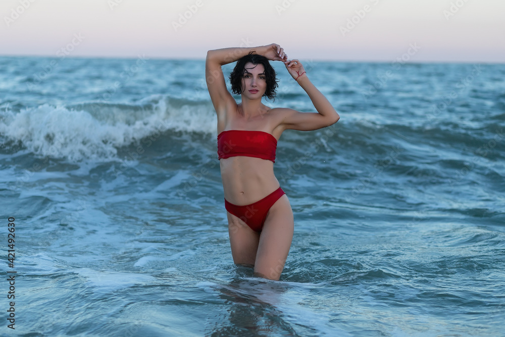 Summertime recreation concept. Beautiful young sexy woman with fit trained slim body wearing red swimwear bikini poses on a sandy beach. Fashion female model near the sea.