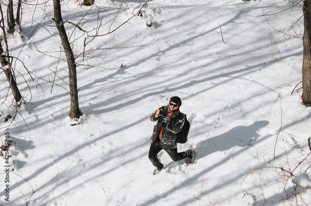 A young male tourist in black warm clothes and a large backpack on his back walks through the snowy forest and takes pictures of nature on a mobile phone.
