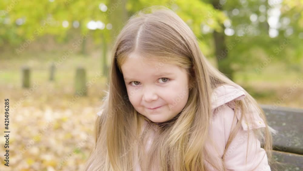 Cute little Caucasian girl smiles shyly at the camera as she sits on a bench in a park - closeup