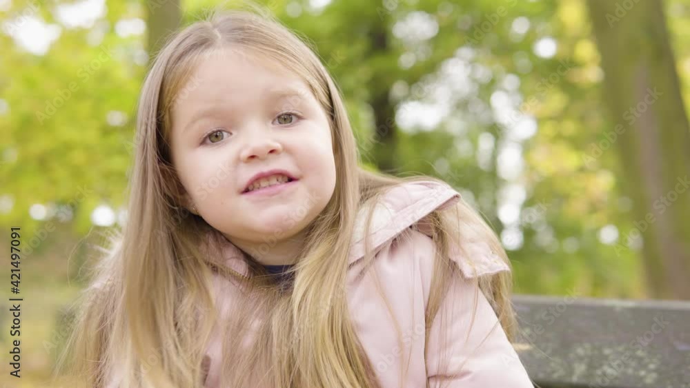 Cute little Caucasian girl dances and smiles at the camera as she sits on a bench in a park - closeup Cute little Caucasian girl dances and smiles at the camera as she sits on a bench in a park - closeup
