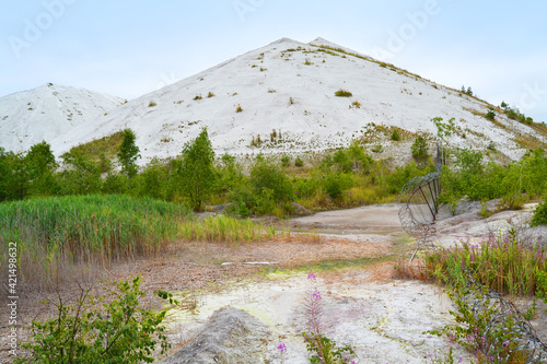 White slag heaps of a Chemical Plant on the background and fallen fence with barbed wire on the right. Phosphogypsum mountains. July 2020, Gomel, Belarus.