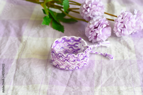 Purple DIY friendship bracelet with unusual braiding on lilac textile next to flowers