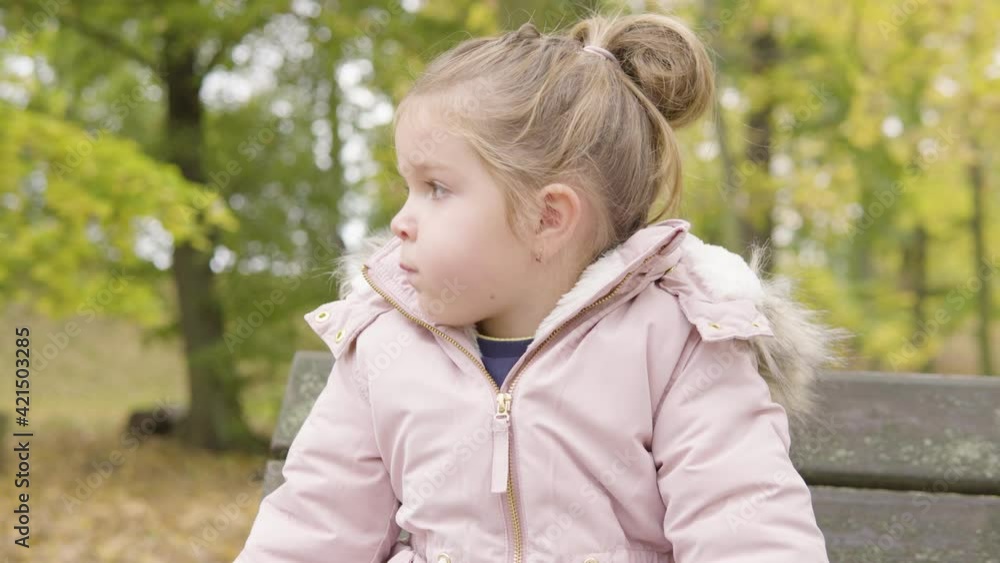 Cute little Caucasian girl looks at something off the camera and eats something as she sits on a bench in a park - closeup Cute little Caucasian girl looks at something off the camera and eats something as she sits on a bench in a park - closeup