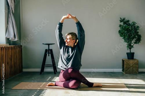 Fototapeta Naklejka Na Ścianę i Meble -  An attractive middle-aged woman does gymnastic exercises at home on a sports mat. Woman doing yoga in the morning in sunlight indoors. Healthy sports lifestyle, sportswear for activity.