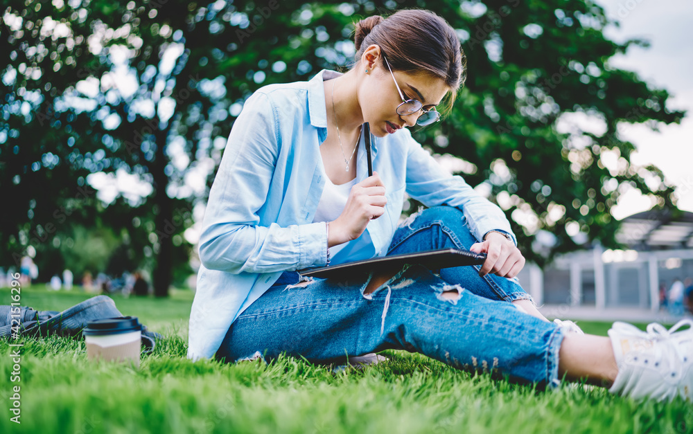 Smart female student in optical eyeglasses reading compendium preparing to school exams sitting at grass and thinking, contemplative hipster girl in spectacles thinking about education while studying Smart female student in optical eyeglasses reading compendium preparing to school exams sitting at grass and thinking, contemplative hipster girl in spectacles thinking about education while studying