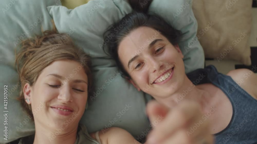 High angle shot of young lesbian couple lying together on bed at home, smiling and talking while holding arms outstretched and touching hands