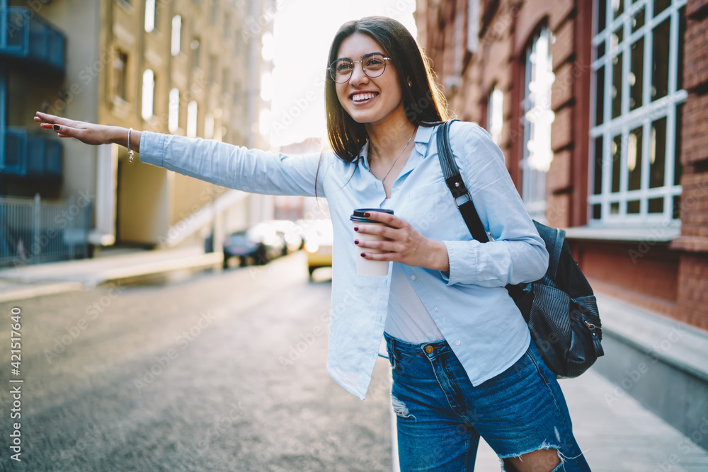 Happy cheerful woman in glasses for vision protection gesturing while standing near road for stopping taxi, funny Caucasian hipster girl with coffee to go smiling while catch public transport
