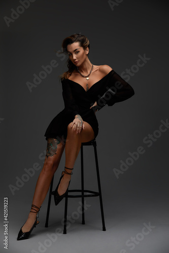Young attractive brunette sexy woman sits on chair in erotic black cocktail dress. Gray background