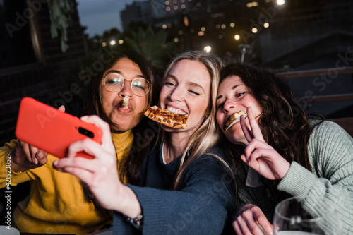 Multiracial girls having fun together outdoors in restaurant while taking selfie with smartphone - Main focus on center girl face