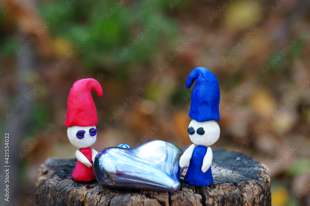 Two plasticine dwarfs in the forest on a colored background. Next to it is a toy heart.