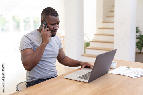 African american man working from home sitting at table using laptop and talking on smartphone,