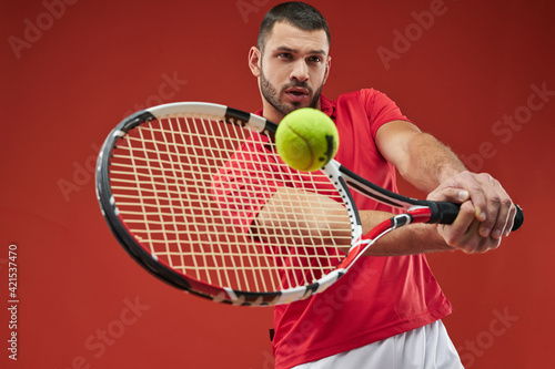 Seriously strong athlete male in red shirt playing tennis isolated on red background © Friends Stock