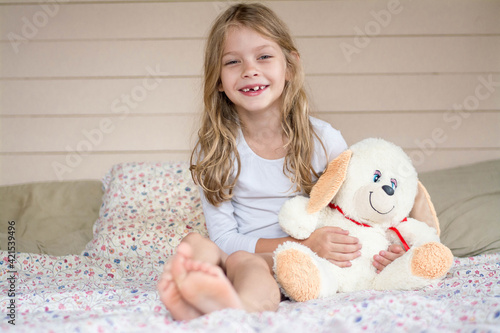 A girl with a plush hare sits on the bed and smiles.