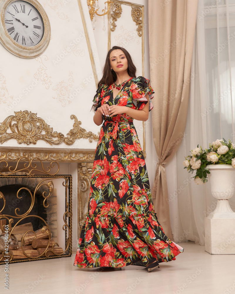 Tall slim cheerful fashion female model in colorful red dress with floral print standing in bright interior