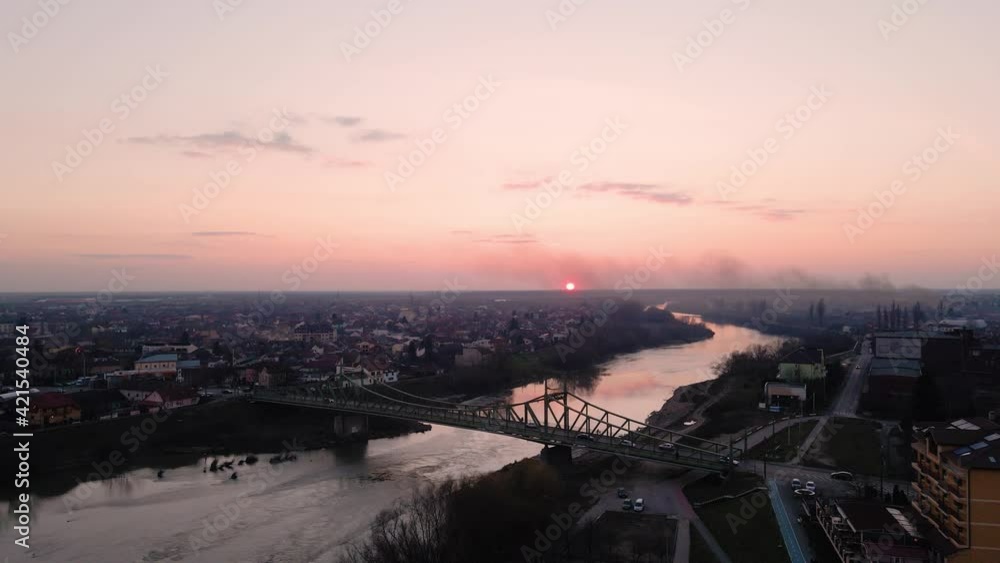 Aerial video of a cityscape with a river and a spanning bridge at sunset. Drone is flying towards the bridge with camera tilting down slightly. Drone footage of cityscape with a river at sunset. Aerial video of a cityscape with a river and a spanning bridge at sunset. Drone is flying towards the bridge with camera tilting down slightly. Drone footage of cityscape with a river at sunset.