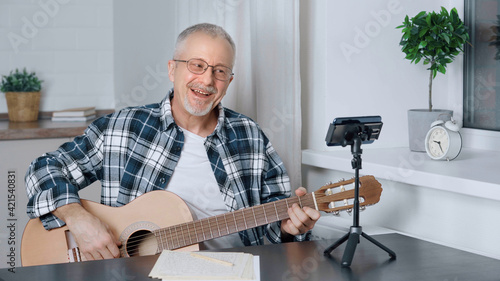 An elderly male blogger teaches guitar playing in an online lesson at home.