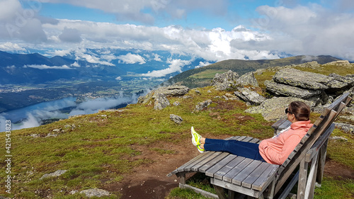 A woman in orange hoodie sitting on a wooden bench with a view on the Alpine valley from the top of Granattor in Austrian Alps. The valley is shrouded in fog. Many mountain poking above the clouds photo
