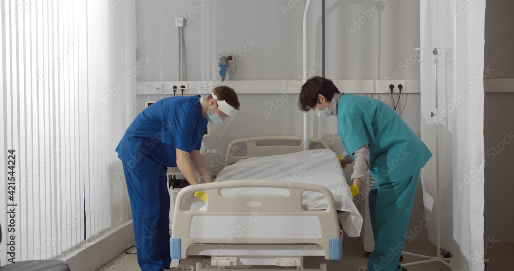 Medical workers in safety mask and gloves changing bed sheets in hospital room Medical workers in safety mask and gloves changing bed sheets in hospital room