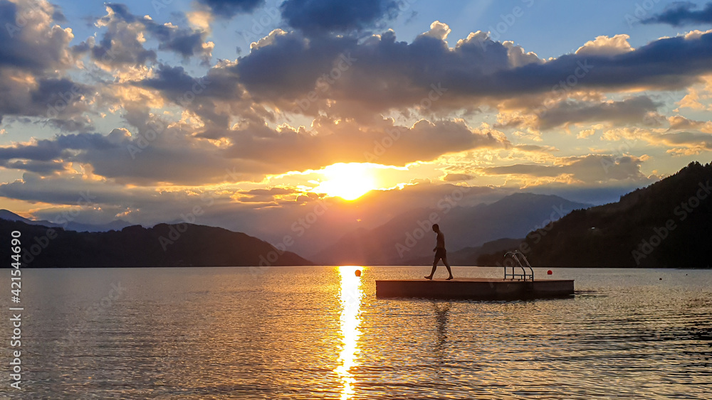 A man running along wooden platform drifting on the MIllstaetter lake during the sunset. Jumping into deep water. The sun sets behind high Alps. Calm surface of the lake reflects the orange sky. Fun