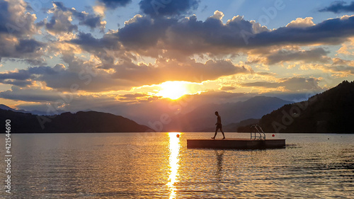 A man running along wooden platform drifting on the MIllstaetter lake during the sunset. Jumping into deep water. The sun sets behind high Alps. Calm surface of the lake reflects the orange sky. Fun photo