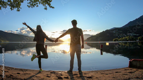 A couple standing at the shore of Millstaetter lake and holding hands during the sunset. The sun sets behind high Alps. Calm surface of the lake reflects the orange sky. They are having fun. Day end