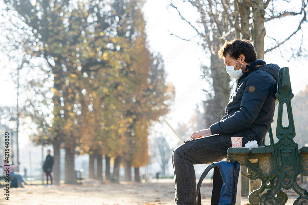 Side view of man wearing protective face mask using laptop in public park Side view of man wearing protective face mask using laptop in public park