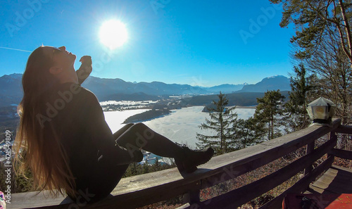 A woman in black sitting at the railing of a viewing platform and flipping her long, brown hair. There is a frozen Faaker lake underneath, surrounded by high Austrian Alps. The woman enjoys sunny day