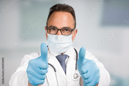 male doctor in lab coat with face mask and stethoscope shows thumbs up